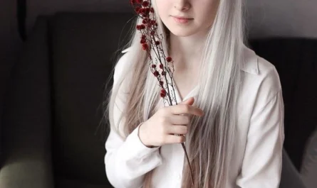 A young girl with long white hair and different-colored eyes sitting at a café table, holding a branch with red flowers.