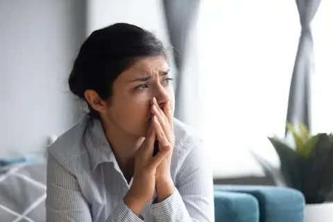 Worried young woman sitting indoors, holding her hands near her face, looking anxious or stressed.
