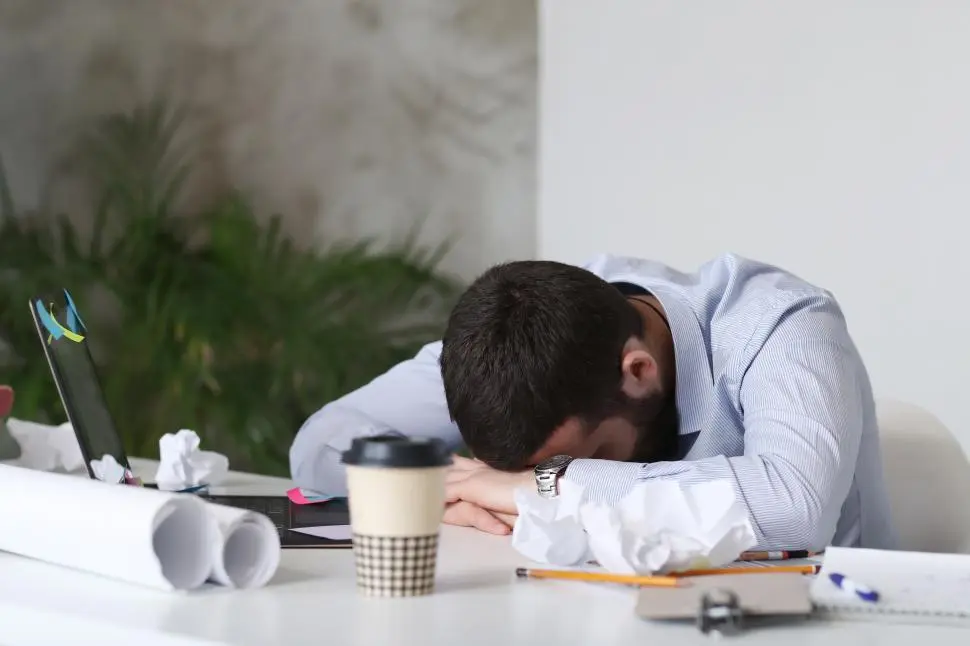 Tired office worker resting his head on a desk surrounded by paperwork, laptop, and coffee cup.