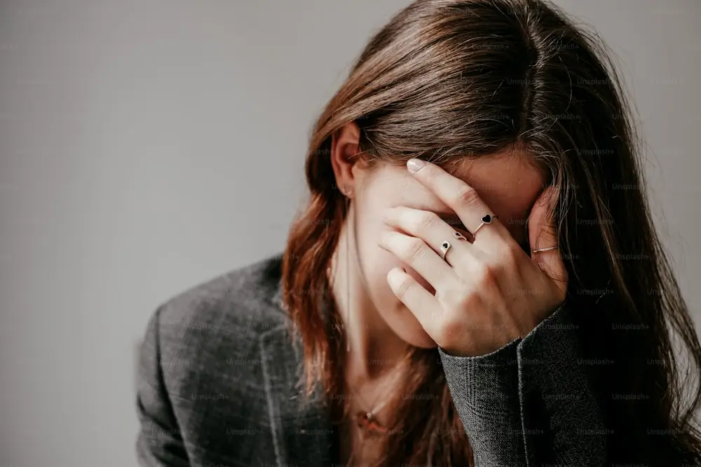 Young woman in a gray blazer covering her face with her hand, expressing emotional stress or fatigue.